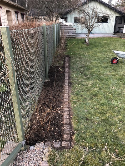 Nygrävd rabatt längs staket med uppradade trottoarstenar och en skottkärra i bakgrunden.