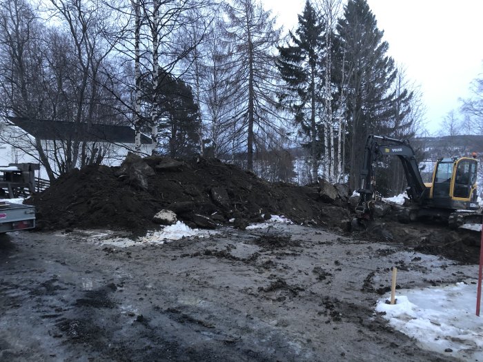 Grävmaskin och jordhög på lerblandad byggarbetsplats med snörester och träd i bakgrunden.