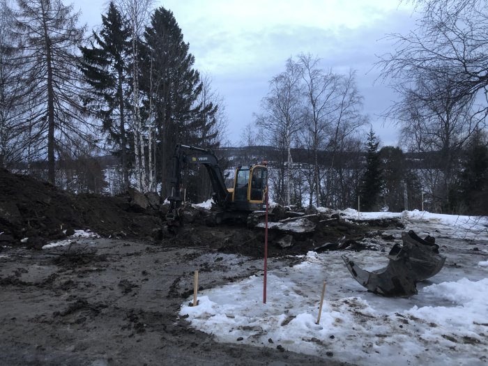 Grävmaskin på lerig byggarbetsplats med smältande snö och avgrävningsarbete i gång.