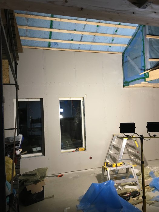 Nästan färdigbyggd vägg med gipsskivor och ett fönster, stegar och byggmaterial syns.