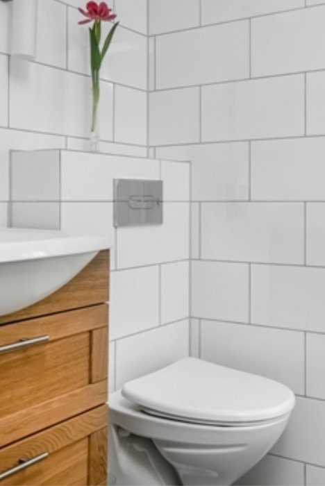 Badrum med vit toalett och handfat, träskåp och vita kakelväggar med en blomma i vas.