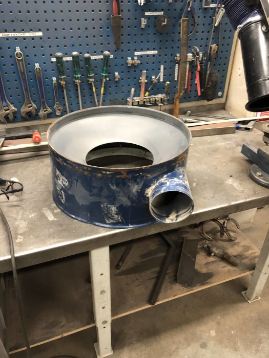 En modifierad trumma från en spånsug med påsvetsad metallbit på en arbetsbänk i en verkstad.