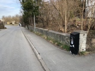 Soptunna vid vägkant med stödmur och nakna träd i bakgrunden på en grusig gata.