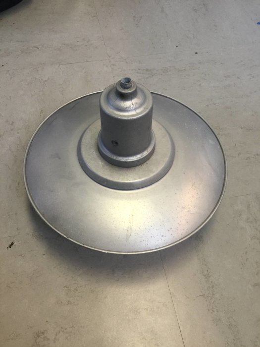 En gammal metall stalllampa på ca 40 cm i diameter ligger på ett grått golv, avsedd för trådom.