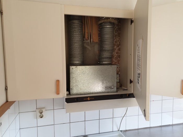 Ventilationsutrymme ovanför köksfläkt med öppen skåpdörr och synliga ventilationsslangar.