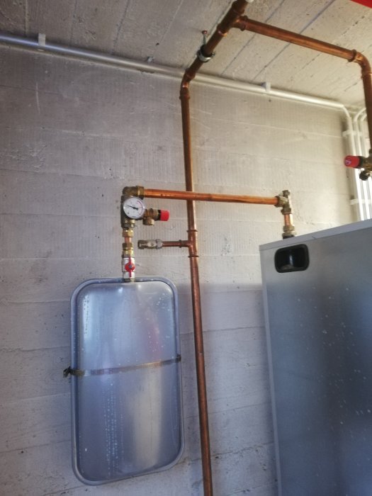 Mätare monterad på vägg med kopparledningar och plåtfärgat kärl nedanför i en installation.