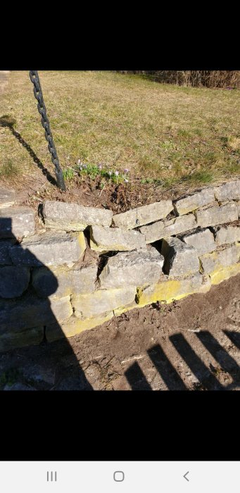 Skadad stenmur vid trädgårdens entré med flera lösa och förskjutna stenblock mot en sluttande gräsmatta.