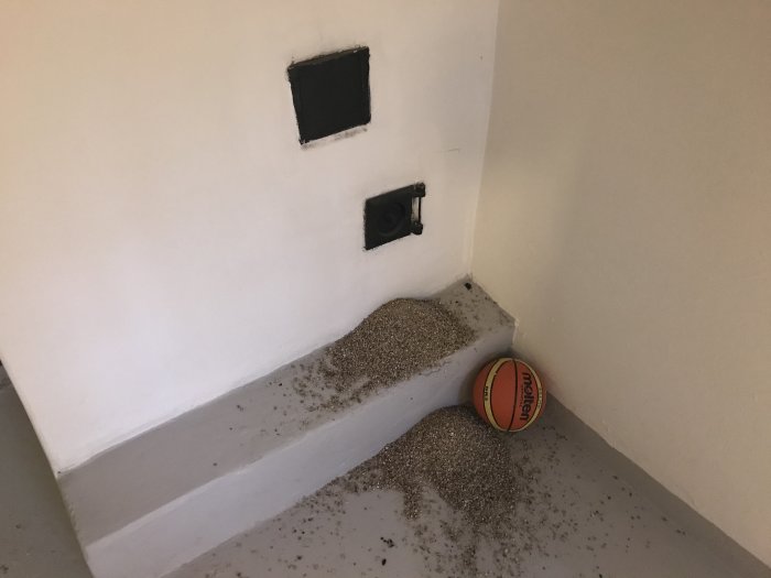 Hörn av rum med lucka i murstock och utspridd hög av småkornigt grus på golvet bredvid en basketboll.