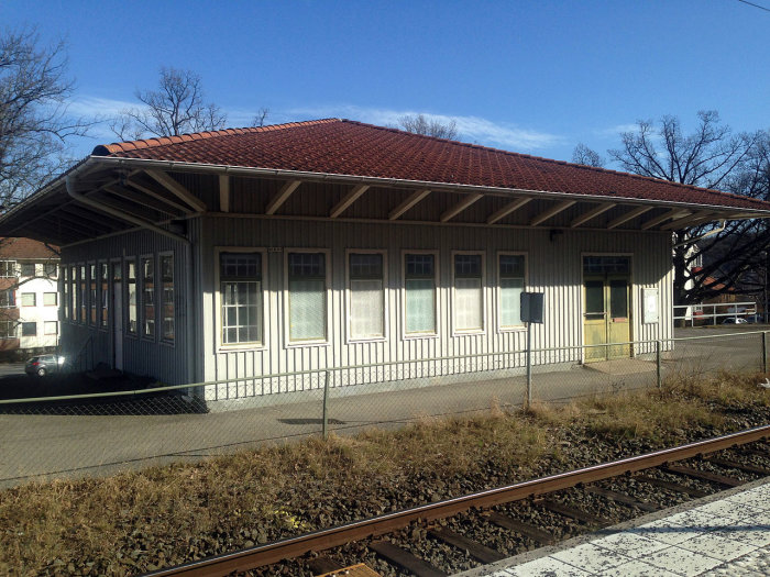 Bild av den traditionella stationsbyggnaden i Huskvarna vid järnvägsspår under solig himmel.