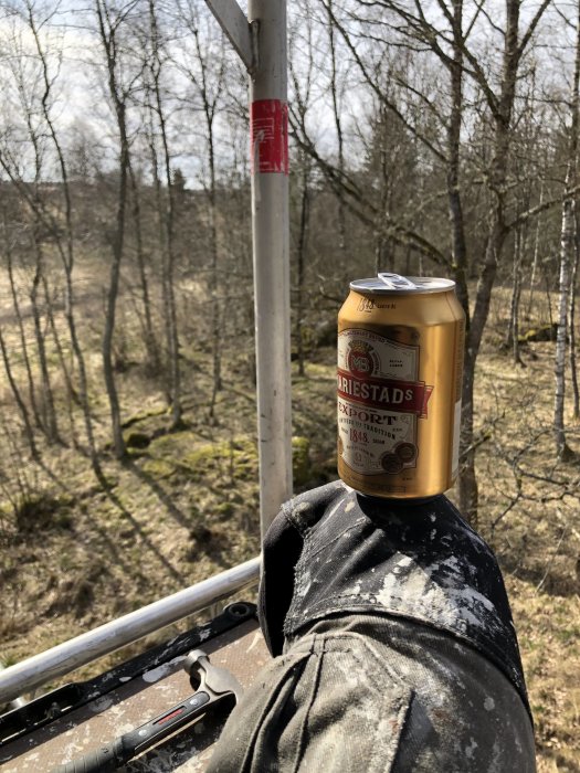 Burk öl balanserad på en byggnadsarbetares knä högt upp på byggnadsställning med skog i bakgrunden.