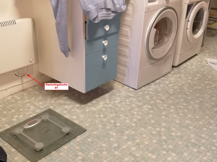 Tvättstuga med tvättmaskin, torktumlare, skåp och markerad plats för rensningslucka på golvet.