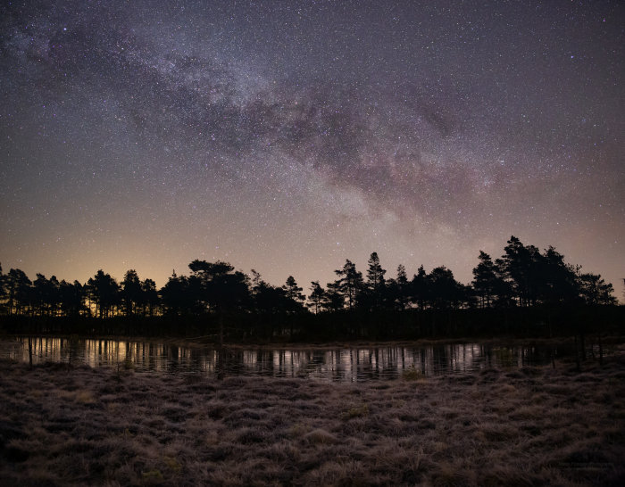 Stjärnklar himmel över frostigt landskap och silhuett av träd mot sjö vid gryning.