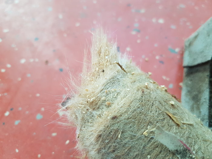 Misstänkt rörisolering med fibrösa material och små partiklar mot röd bakgrund.