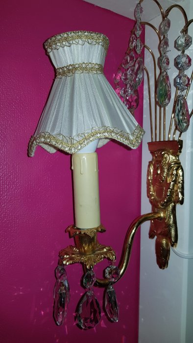 Vintage vägglampa med vit ljuskälla, gyllene detaljer och hängande kristaller mot en rosa vägg, förmodad äldre modell.