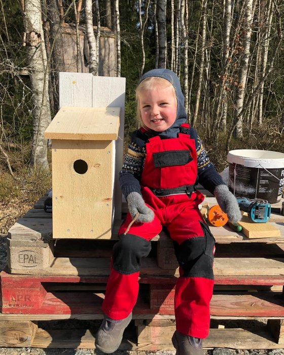 Leende barn i snickarbälte sitter bredvid nybyggd fågelholk utomhus.