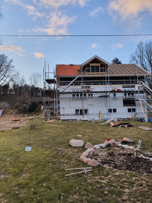 Hus under renovering med nytt tegeltak i arbete, byggnadsställning och byggmaterial spridda på gräset.