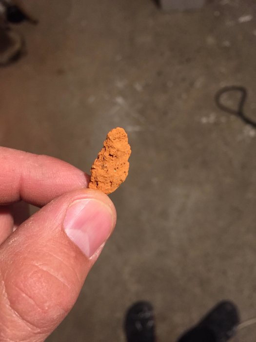 Hand håller ett finkornigt rött dammparti mot en suddig bakgrund av ett källargolv.