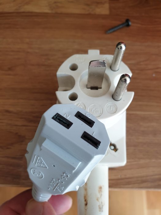 Hand håller en vit elektrisk adapter med en äldre 230V stickpropp i förgrunden, framför ett vägguttag för Schuko-stickkontakter.