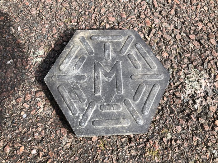 Svart klinkerplatta med präglat mönster och bokstaven M, liggande på grov asfalt.