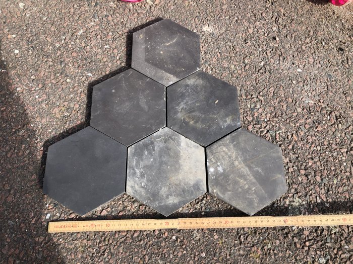 Svarta hexagonala klinkerplattor bredvid en linjal på grusunderlag som visar dess storlek.