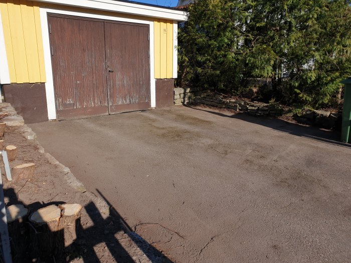 Nyskottad garageuppfart med trädstubbar i förgrunden och en gammal garageport mot en gul vägg.