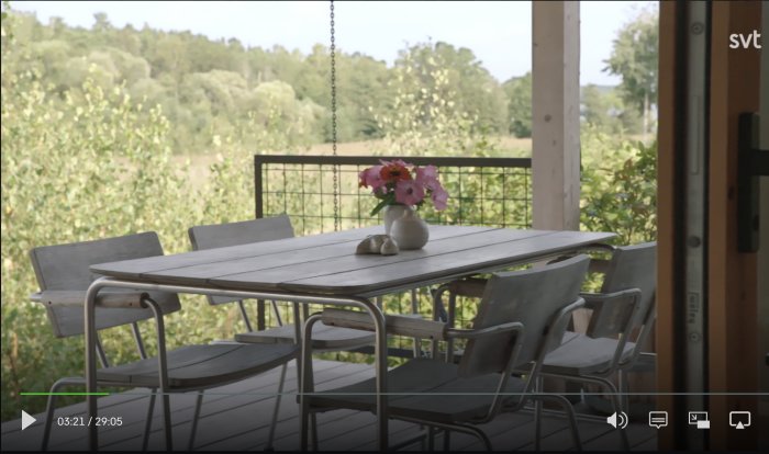 Utemöbler med bord och stolar på en veranda, omgiven av grönska, med en vas blommor på bordet.