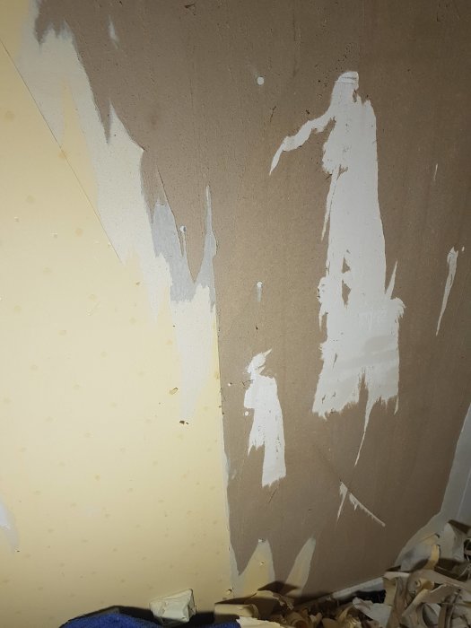 Vägg under renovering med avlägsnade tapetlager som avslöjar brun spackel och vit underliggande yta.