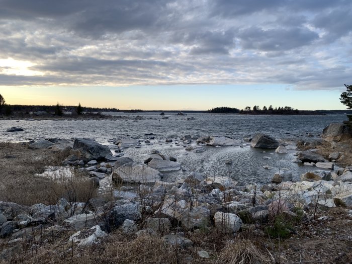 Kustlandskap vid solnedgång med stenar och is vid vattenbryn, molnig himmel i bakgrunden.