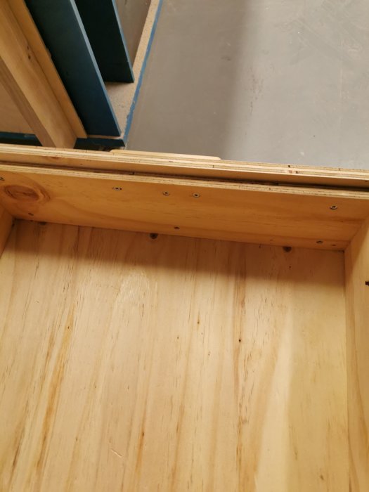 Hörn av hemgjord plywoodlåda med fräst fals och skruvade detaljer synliga samt en lackad björklist.