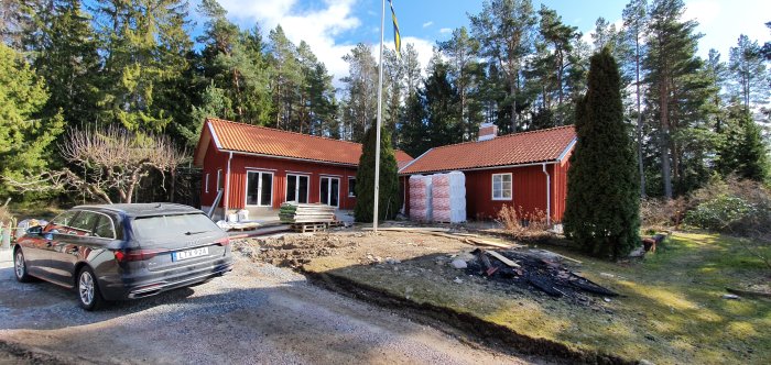 Röd stuga med nytt tak och nockpannor, byggmaterial och en bil framför, under blå himmel.