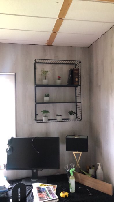 Industriell vägghylla med tre hyllplan ovanför ett skrivbord, inrett med växter och dekorativa föremål.