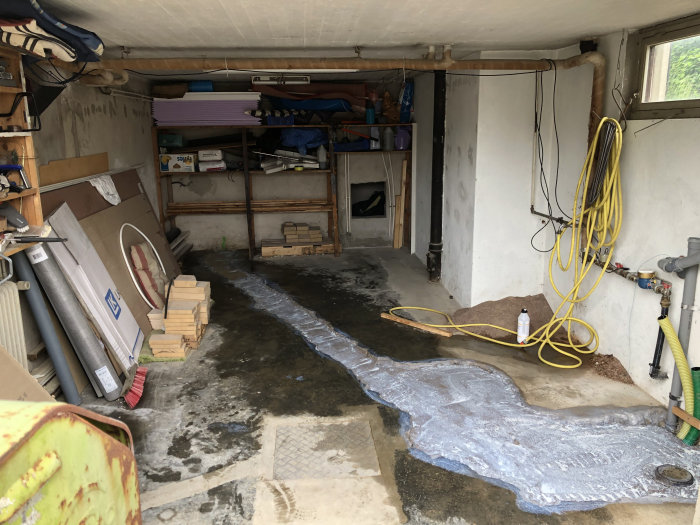 Ett garage med nylagt primer på golv där avlopp dragits, byggmaterial staplas, och en sliten sopborste syns.