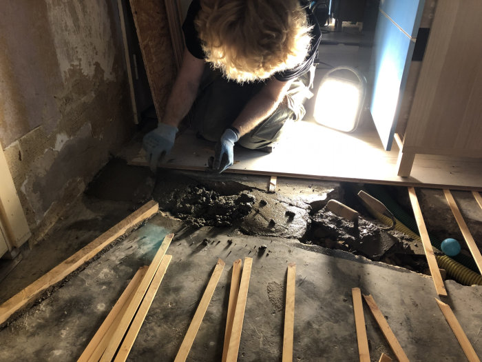 Person reparerar golv i garage med synligt avlopp och träreglar, arbetsbelysning på golvet.