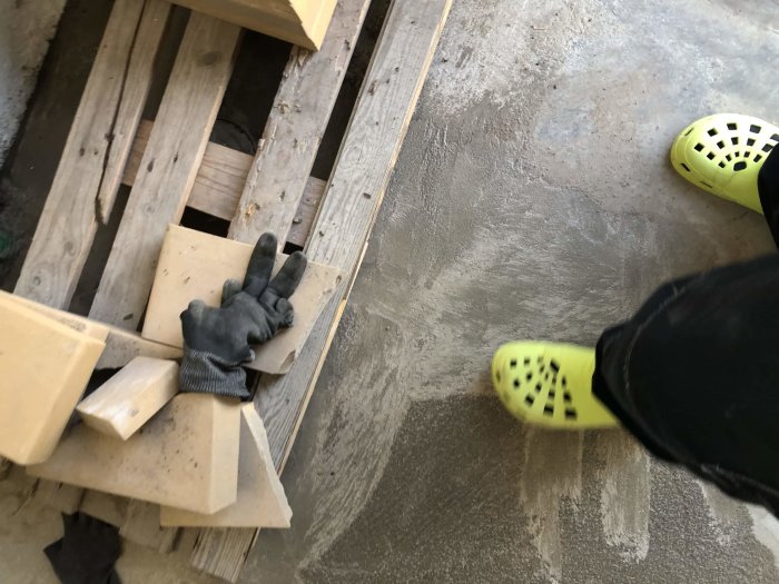 Renoveringsarbete med nylagt betonggolv, träplankor och arbetshandske, syn av person i gula skor.