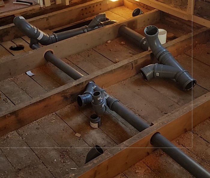 Upprivet golv med exponerade träbjälkar och nydragna avloppsrör i ett badrums renoveringsprojekt.