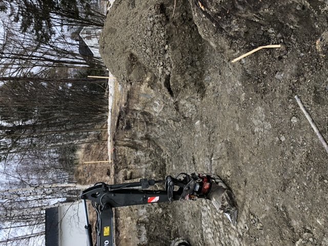 En stor utgrävd grop med en grävmaskin i framkant, markerad för vidare grävning, i en trädgård.