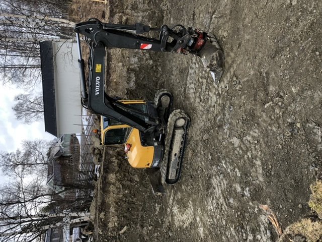 Grävmaskin i färd med att gräva en stor grop på en byggarbetsplats, omgiven av grus och jord.