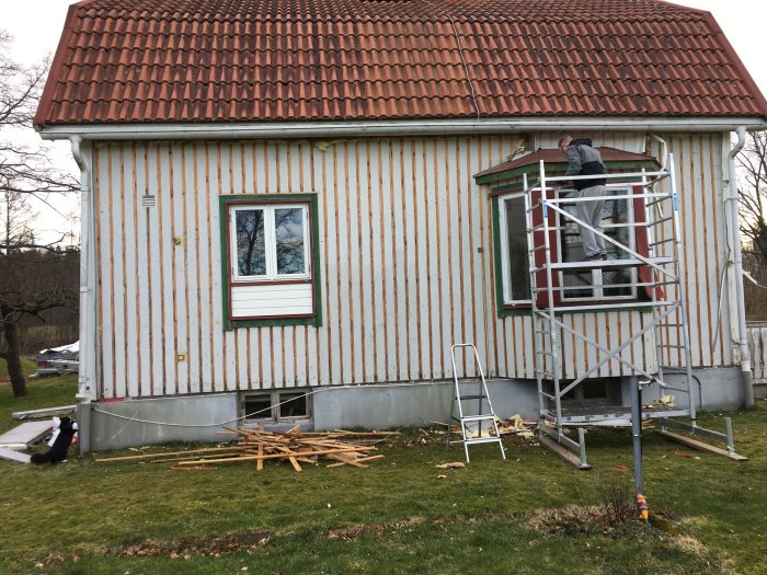 Person på byggnadsställning renoverar fasad på tvåvånings trähus med tegeltak.