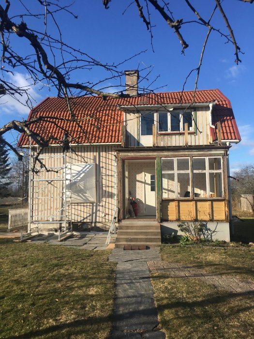 Traditionellt tvåvåningshus under renovering med ställningar och oskyddad isolering på ena väggen, blå himmel i bakgrunden.