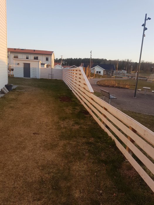 Nybyggt trästaket längs en gräsmatta med lekpark i bakgrunden, staketet lutat i väntan på stabilisering.