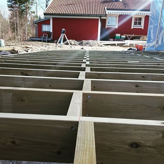 Träbjälklag med korsande kottlingar framför en röd stuga under konstruktion.