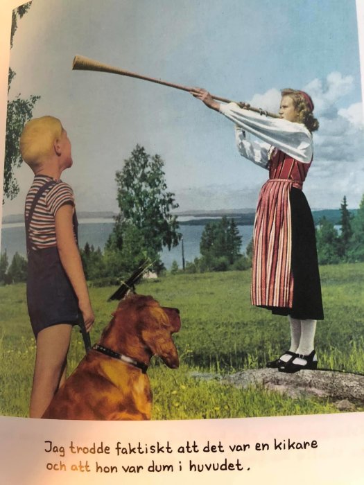 Kvinna i traditionell klädsel blåser i ett långt horn medan en pojke och en hund tittar på, mot bakgrund av en sjö och skog.