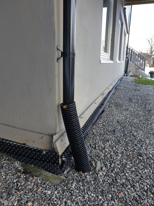 Stuprör leder regnvatten direkt i ett svart dräneringsrör vid en husgrund från 1940-talet.