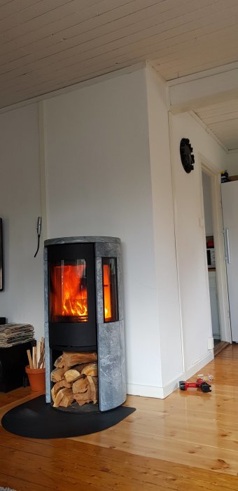 Nyinstallerad kamin med brinnande eld i en hörna av ett rum, synlig spricka i väggen nära taket, ved staplad undertill.
