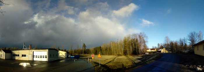 Panoramavy över industribyggnader, en skogskantad väg och ett bostadshus mot en dramatisk himmel.