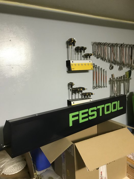 Ljusskylt med texten "FESTOOL" på en verkstads hylla ovanför en kartong.