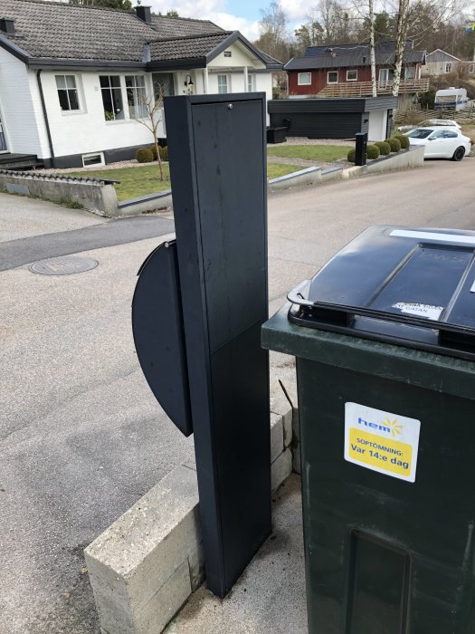 Solblekt svart brevlåda vid husentré med en öppen lucka och sophämtningsschema på soptunna.