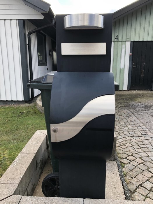 Svart brevlåda med solblekt färg och rostfritt stål framför hus och stenlagd gång.