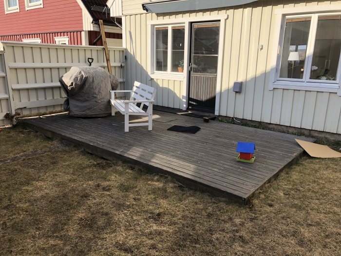 Gammalt träträdäck nära huset med en vit stol, täckt grill och leksaker, indikerar renoveringsbehov.