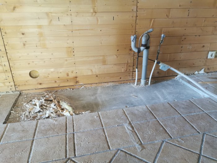 Renoverat kök med riven del av klinkergolv och synliga rörinstallationer mot trävägg.